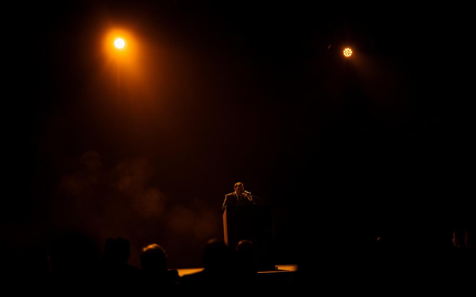 Speaker at a lectern under a single spotlight on a darkened conference stage